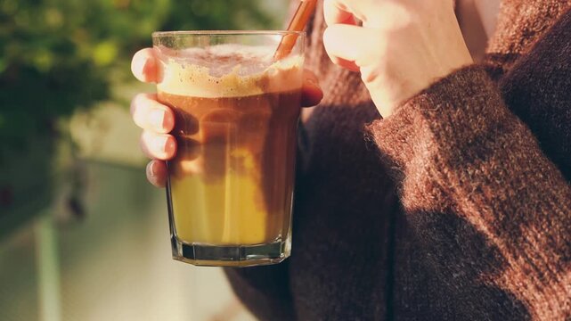 Woman stirring a homemade pumpkin spice latte with a straw, creating swirling layers inside the glass. Cozy autumn lifestyle footage in warm golden sunlight with a knitted cardigan