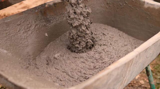 Wet gray concrete pours into metal wheelbarrow at outdoor construction worksite, building thick lumpy mound with rough damp texture under warm daylight in overhead view.