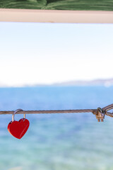 Red heart shaped love padlock attached to a steel cable above the sea with a plain reverse side and softly blurred blue coastal water creating a romantic travel themed close up © Duncan