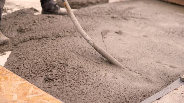 Fresh wet concrete spreads over construction formwork as hose crosses diagonally, showing coarse aggregate texture, smoother leveling, and partial worker boots at active building site.
