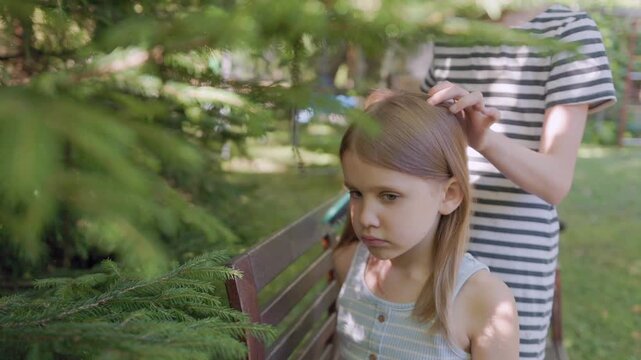 Young girl sits outdoors on wooden bench while older sister carefully combs her blonde hair with brush. Calming sister hair grooming routine continues peacefully under garden trees.