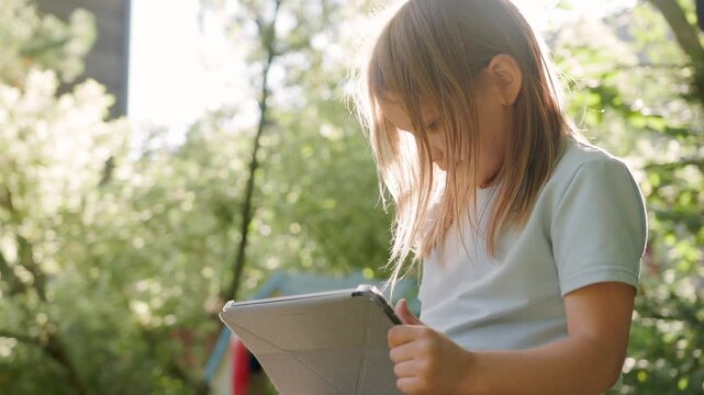 Young girl plays digital tablet outdoors in sunny park setting, focused on portable screen with intense attention while sitting outside under bright sunlight during playful summer afternoon.