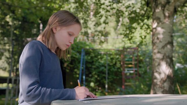 Child sketching outside sits by wooden table as sister walks over. Creative young girl draws with pencil on paper in sunny backyard while sibling observes artwork curiously.