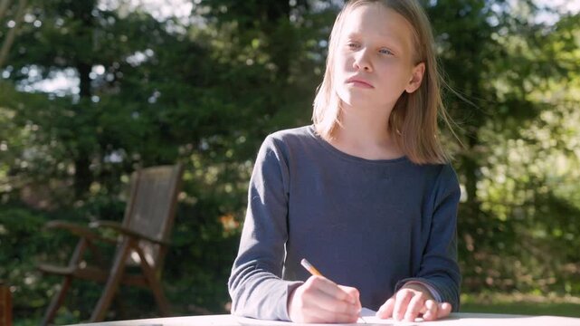 Focused child sketches on paper using pencil during outdoor kids art therapy. Thoughtful young girl sits at table in backyard garden, engaging in peaceful drawing activity with calm expression.
