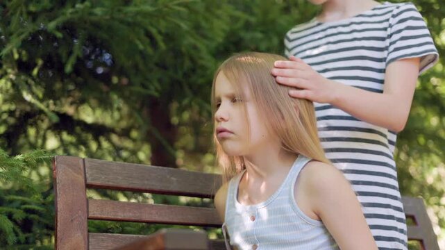 Blonde girl sits outside while sibling brushes long hair with paddle brush. Expression turns from calm to annoyed as gentle brushing continues beside green garden trees on sunny afternoon.