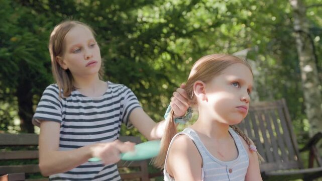 Young girl carefully grooms long hair of her sister using brush in sunny backyard garden. Two Caucasian children sit together outside on bench, sharing gentle sibling care and quiet bonding moment.