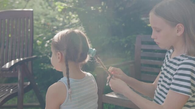 Older sister styles hair of little child in backyard during peaceful family summer routine. Caring girl carefully ties cute braid for relaxed sister seated outside on wooden patio furniture.