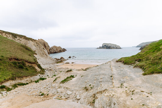 A view of a sandy beach with rock formations and shallow water. Gray clouds are visible in the sky. Signs of the receding tide are evident along the shoreline.