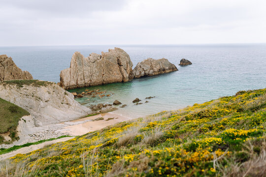 A view of a sandy beach with rock formations and shallow water. Gray clouds are visible in the sky. Signs of the receding tide are evident along the shoreline.