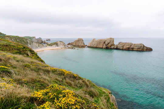 A view of a sandy beach with rock formations and shallow water. Gray clouds are visible in the sky. Signs of the receding tide are evident along the shoreline.