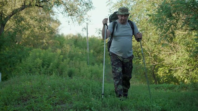 Hiker with backpack and trekking poles walking toward camera through lush woodland meadow wearing camouflage trousers and sun hat in a natural outdoor setting