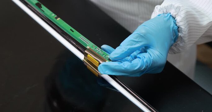 Technician wearing blue gloves installs a flexible circuit board along the edge of a black display panel in an electronics factory.