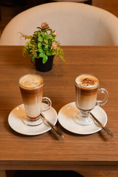 Two cups of cappuccino in tall glass mugs on white saucers with teaspoons, on a wooden table.