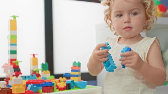 Blonde little girl stacking colorful plastic building blocks and reaching into a tub on a play table inside a bright playroom building; curiosity.