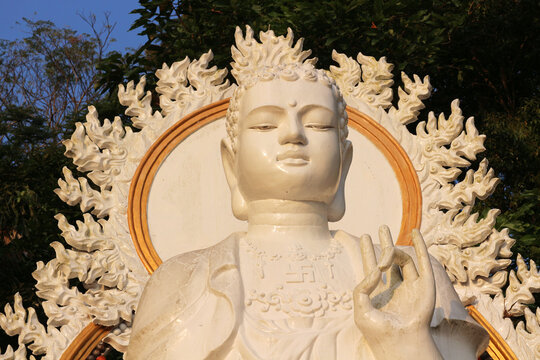 Close up of a serene white Buddha statue with an ornate flame-shaped halo and hand gesture.