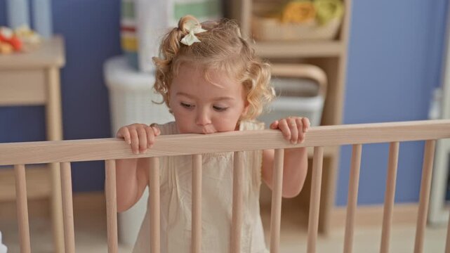 Little blonde girl holds crib railing with both hands while standing in a nursery, peeking over the rail at toys and room; curiosity innocence playtime.