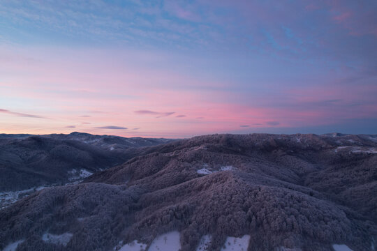 Aerial view of beautiful soft pink sunset sky over winter mountain forest covered in hoarfrost, drone panoramic shot of snowy hills and frozen trees in evening twilight light, carpathians landscape