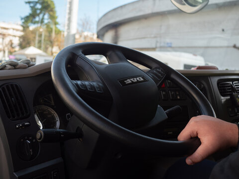 DAF brand logo on the black plastic steering wheel inside a modern commercial heavy duty truck cabin featuring the hand of a professional male driver operating the large vehicle.