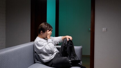 Young man sitting alone on a sofa in a dark room, covering his face with his hands and holding a game controller, expressing defeat and exhaustion after a long gaming session © Aliaksandr