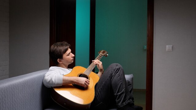 Young man sitting comfortably on a sofa in his home, enjoying a moment of leisure while practicing a musical instrument and creating melodies in a relaxed setting