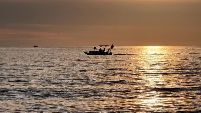 Silhouette of a small fishing boat sailing at sunset, cinematic golden hour scenic ocean sea view.