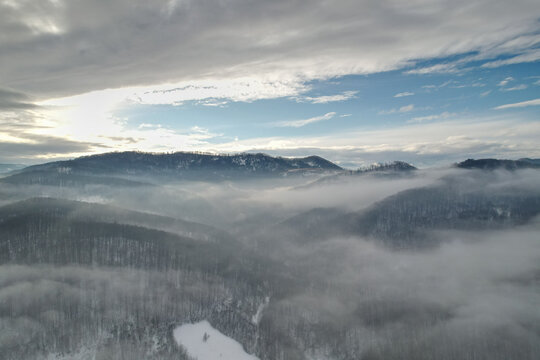 Aerial view of majestic winter mountains covered in snow and low rolling clouds, drone shot of foggy valleys and forest ridges under blue sky with sunlight, beautiful scenic nature landscape
