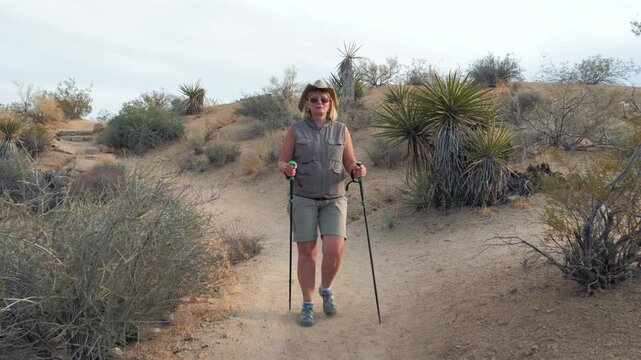 Mature woman with trekking poles walking on sandy trail enjoying an active outdoor lifestyle in dry arid landscape with desert plants and hills during her adventurous vacation. Front view in motion