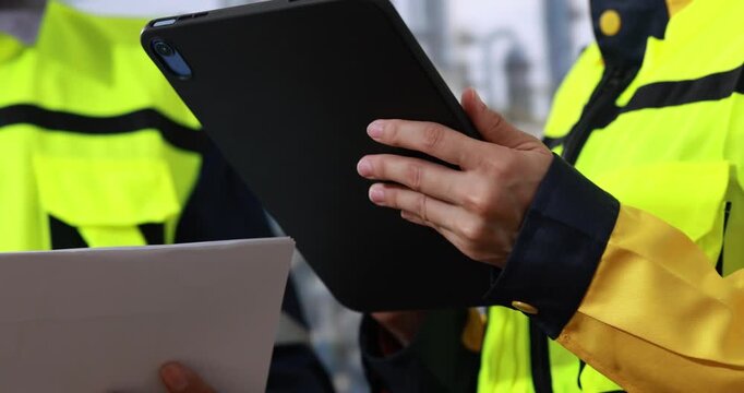 Close-up of safety workers in high visibility jackets using a tablet and clipboard during an outdoor inspection.