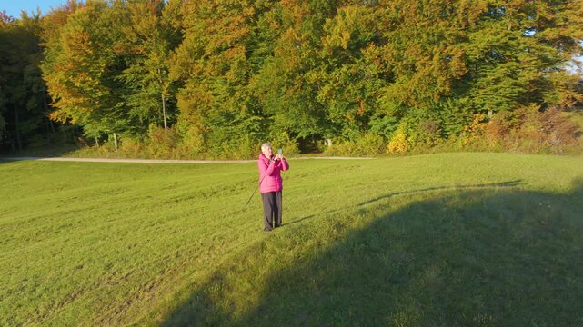 AERIAL: Senior woman pauses after hiking to photograph a colourful autumn landscape with her smartphone. She stands in a sunny meadow with walking poles, surrounded by vibrant forest and golden light.