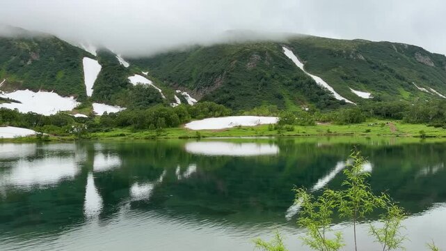Tokhkolach Lake reflects misty Vachkazhets mountain slopes with lingering snowfields in Kamchatka, Russia. Pristine wilderness, mountain scenery, nature tourism, tranquility and remote travel