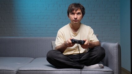 Young man sitting comfortably on a sofa, deeply engrossed in playing a video game using a wired controller, illuminated by the screen's glow in a cozy living room setting © Aliaksandr