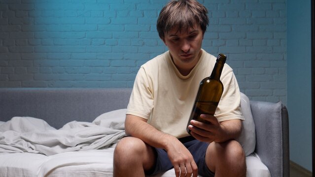 Man with an unhealthy appearance and messy hair sitting on a bed in a messy room, feeling lonely and depressed while holding an empty bottle, symbolizing his addiction and struggle