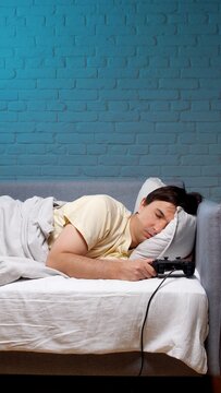 Young man asleep on sofa clutching a wired game controller, exhausted from late night gaming, depicts sleep deprivation, gaming addiction and tech overuse affecting rest patterns