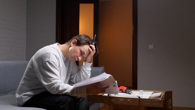 Young man feeling stressed and confused while trying to assemble flat pack furniture, sitting on a sofa and looking at complicated instructions with a screwdriver in his hair
