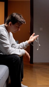 Man holding a tangled knot of wired earphones, showing frustration while sitting on a sofa in a domestic setting, dealing with connection problems and technical issues