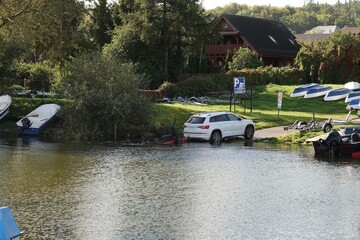 Biały samochód osobowy stoi na slipie przy brzegu jeziora obok przystani z łodziami. © tomo81