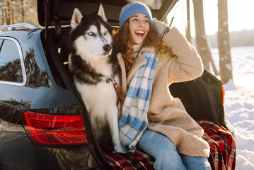 Woman and her husky are relaxing in trunk of car in snowy forest. Happy woman enjoys winter outing with dog, sitting on blanket in car at sunset. Concept of friendship and adventure. Lifestyle. © maxbelchenko