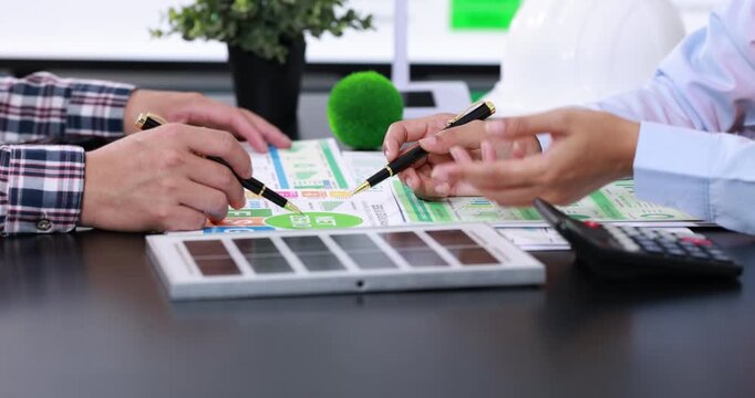 Close-up of two people reviewing a home energy report with a green model tree, solar panel sample and calculator on an office desk.