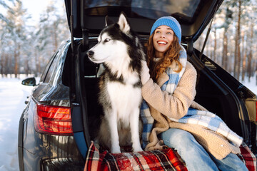 Woman and her husky are relaxing in trunk of car in snowy forest. Happy woman enjoys winter outing with dog, sitting on blanket in car at sunset. Concept of friendship and adventure. Lifestyle. © maxbelchenko