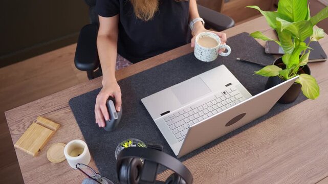 Woman drinks coffee while using laptop and computer mouse, then puts mug down and continues typing. Concept of coffee break and working from home