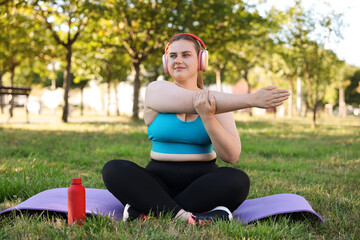 Fototapeta premium Overweight woman with headphones practicing yoga on mat in park