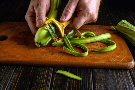 Hands use yellow peeler on zucchini, creating long vegetable ribbons across wooden board. Careful food preparation leaves fresh squash strips ready for home cooked dish, with ribbons spread on board.