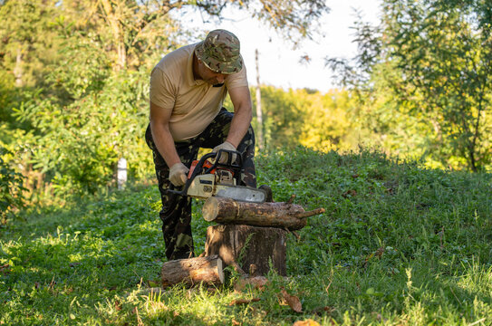 Man operates chainsaw against log resting on stump in leafy woodland. Concentrated lumber worker grips tool with gloved hands, wearing camouflage clothing as cut timber lies among grass and plants.