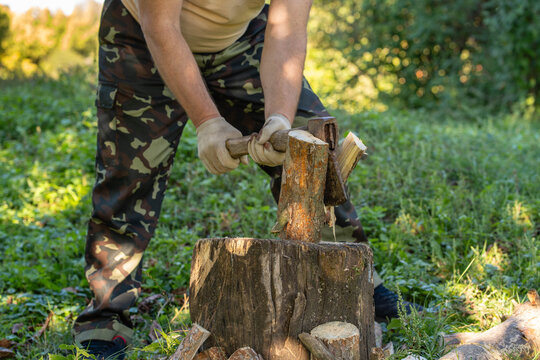 Man holds axe against piece of wood balanced on chopping block, splitting logs in green woodland setting. Gloved worker wears camouflage clothing for outdoor firewood preparation, with axe set