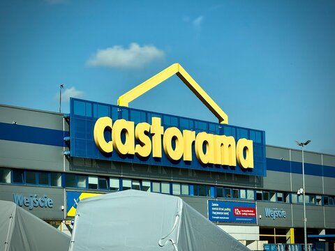 Low-angle view of a Castorama DIY store entrance facade featuring a large blue sign with a yellow logo and house icon under a clear sky, with white promotional tents in the foreground.