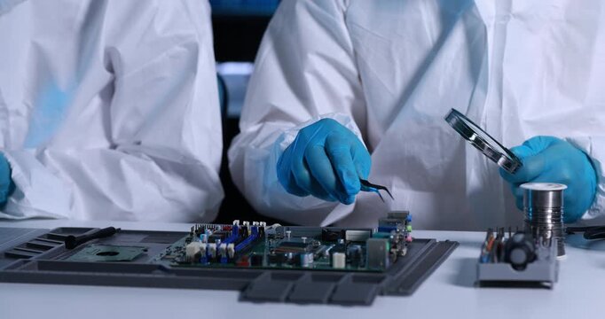 Close-up of technicians in protective coveralls using tweezers and a soldering iron to repair an electronic circuit board in a clean workshop.