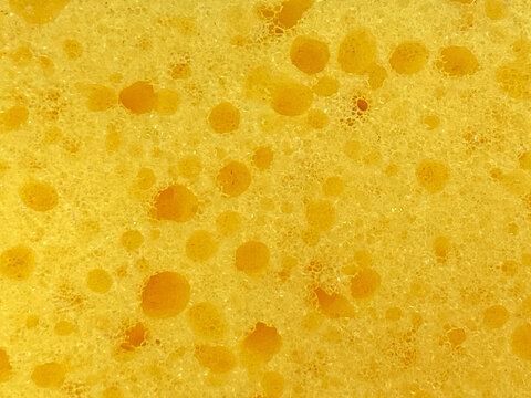 A close-up view of the porous texture of a yellow cleaning sponge filled with irregular holes and fine synthetic cells.