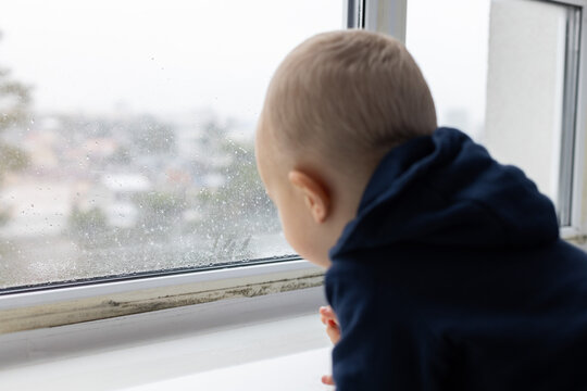 Little child looking through the window with water drops condensate and black spots of mouldy fungus on it. Selective focus.