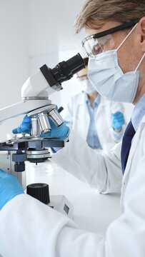 Masked male scientist analyzing sample under microscope, colleague researching nearby in sterile medical laboratory setting. Medicine, healthcare and science concept