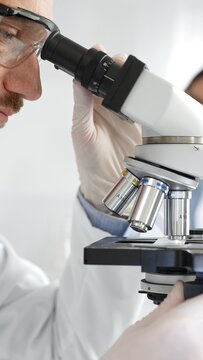 Close up of male researcher examining sample through microscope, wearing safety eyewear in sterile laboratory setting, surrounded by scientific glassware. Medicine, healthcare and science concept